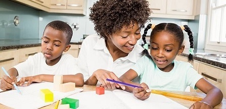 A Black family wearing white t-shirts sits together at a table in a modern-looking kitchen. The adult in the middle has short curly hair and is smiling at the child to her left, whose hair is in braided pigtails. The adult is pointing at the paper they're both looking at, and the child holds a purple writing utensil. The child on the other side of the adult has short cropped hair and is looking down and writing on a piece of paper. There are colorful blocks on the table.