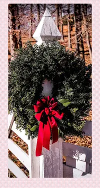 A photo of a wreath made of pine boughs and a red bow on a white wooden fence