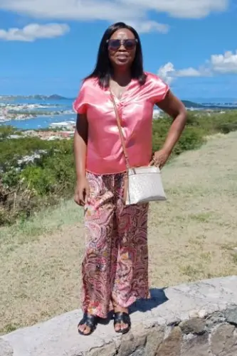 The
  image
  shows
  a
  person
  wearing
  a
  pink
  blouse,
  patterned
  pants,
  and
  a
  clutch,
  standing
  on
  a
  grassy
  area
  with
  a
  scenic,
  partly
  cloudy
  sky
  and
  distant
  mountains
  in
  the
  background.
  AI-generated
  content
  may
  be
  incorrect.