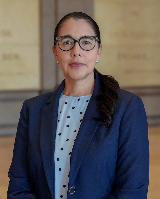 Headshot photo: Dr. Karen Lozano faces the camera with a neutral professional expression, wearing dark-rimmed glasses and a dark blazer. She has long dark hair, parted down the middle and pulled back.